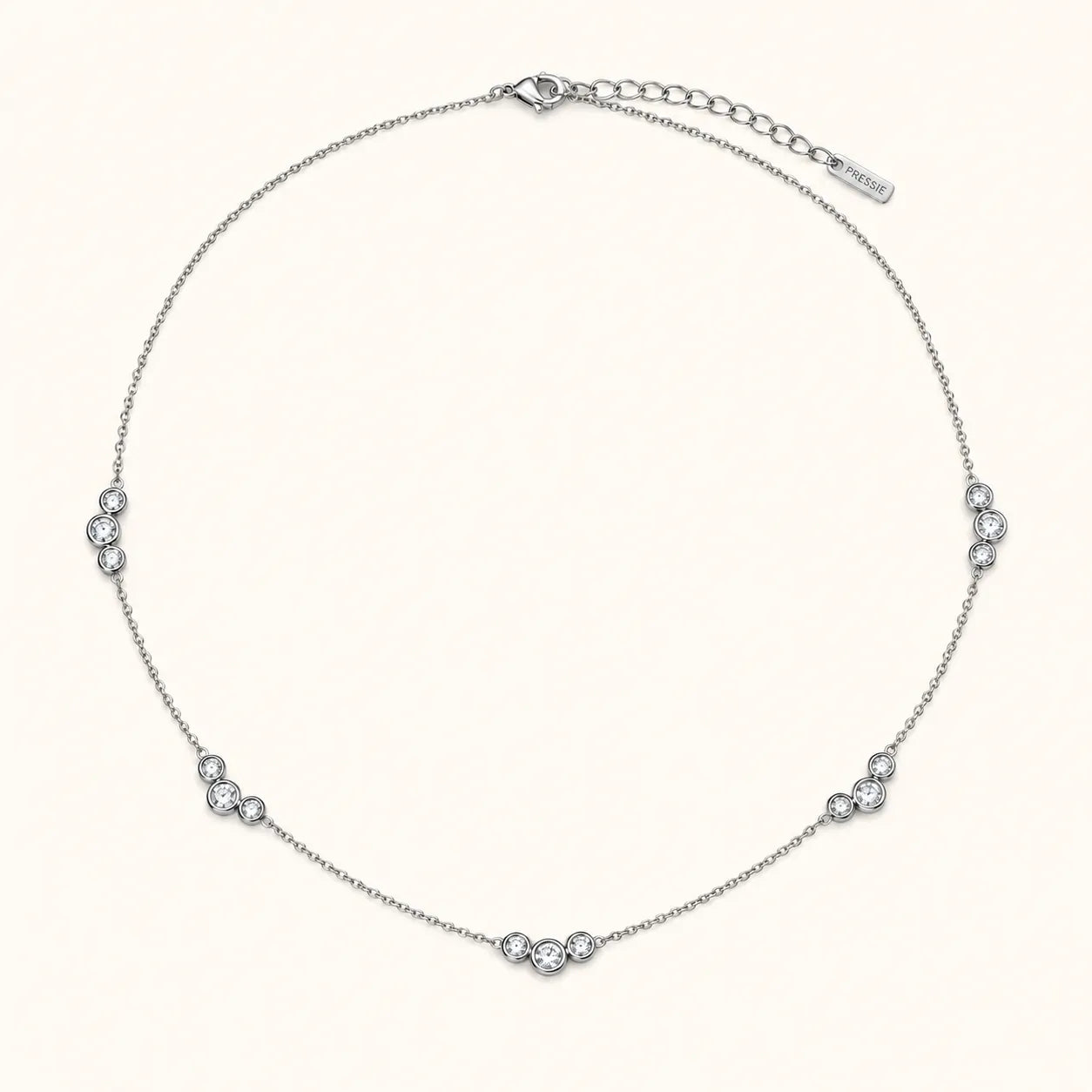 A delicate silver chain necklace with five small clusters of clear round-cut stones spaced evenly along the band, displayed on a light cream background. A brand tag is visible on the extender chain.